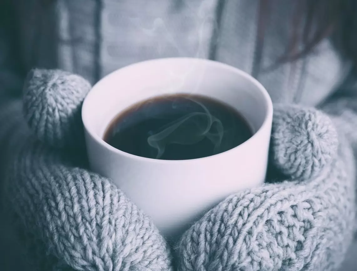 A Person Wearing Mittens Holds A Cup Of Coffee, Surrounded By Two Rocks In The Background.