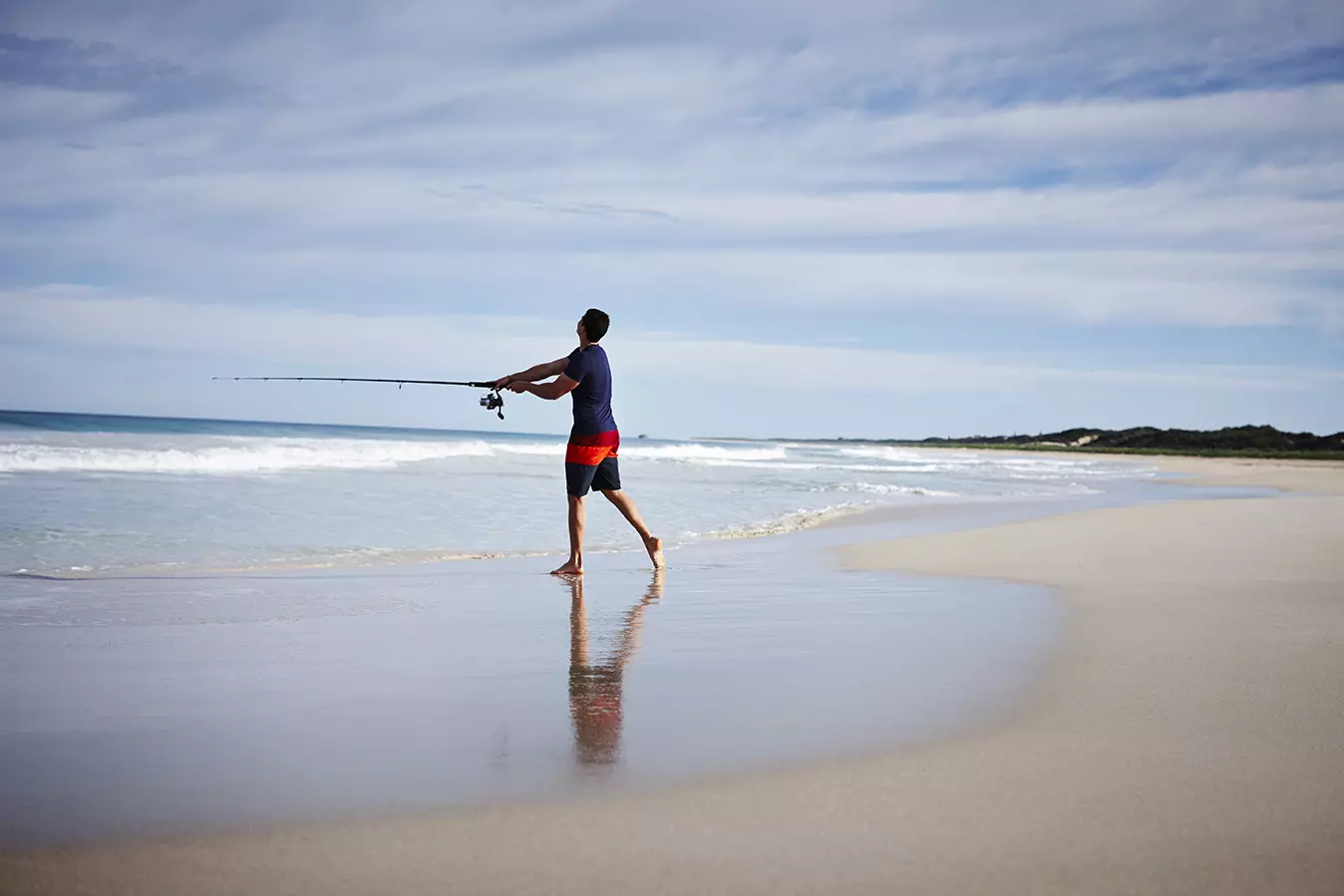 A man fishing on the beach with atlantist two rocks visible in the distance.